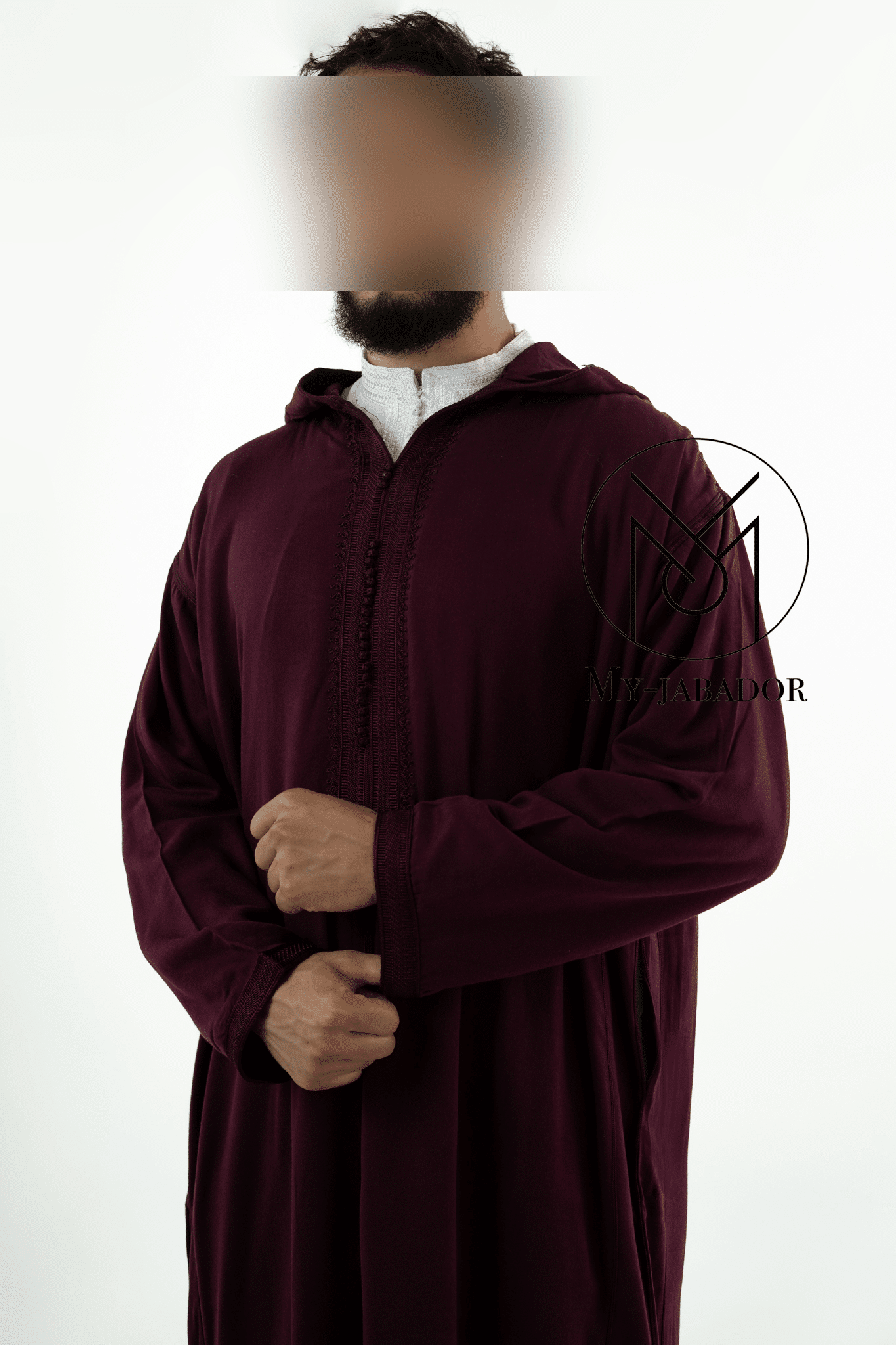 Un homme debout avec élégance, portant une Djellaba d'un riche bordeaux. La tenue fluide met en valeur des broderies délicates le long des manches et du devant. La capuche drapée ajoute une touche de mystère. Une silhouette majestueuse émerge, alliant tradition et élégance dans un bordeaux profond